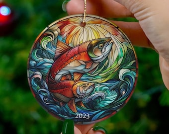 May include: A round ceramic ornament with a stained glass design of two salmon swimming in a blue and green ocean. The ornament has a gold string for hanging and the year 2023 printed on the bottom.