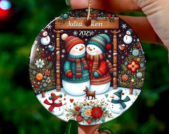Puede incluir: Adorno navideño redondo con dos muñecos de nieve vestidos de invierno, abrazándose bajo un arco de madera. El adorno tiene los nombres "Julia" y "Ken" con el año "2025". El diseño incluye nieve, árboles y detalles florales.