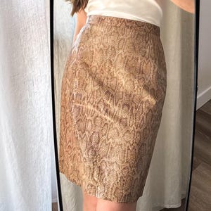 May include: A snakeskin-patterned pencil skirt in brown and tan hues. The skirt, reaching the knee, is made of a smooth material and has a fitted shape. The model is wearing a white t-shirt.