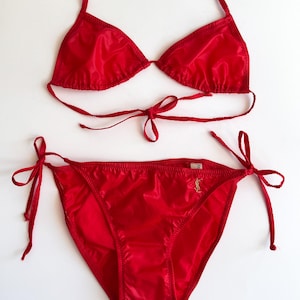 May include: A red bikini set with a halter top and tie-side bottoms. The shiny fabric reflects light, and the top and bottom have tie closures. The bikini is a solid, vibrant red color.