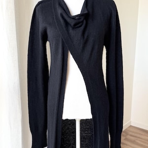 May include: A long, black cardigan with a draped neckline and a single button closure. The open front design reveals a white garment underneath. The sleeves are long, and the cardigan is displayed on a mannequin.