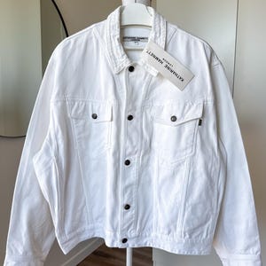 Peut inclure: Veste en jean blanche avec des boutons foncés et deux poches poitrine. La veste a un col classique et une étiquette avec l'inscription "KATHARINE HAMNETT LONDON". La veste est suspendue à un cintre blanc.