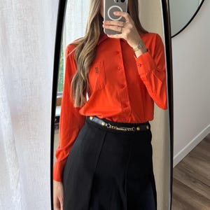 May include: An orange long-sleeve button-down shirt with a small logo on the chest pocket, paired with a black pleated skirt and a black belt with gold chain detailing. The outfit is reflected in a large mirror.
