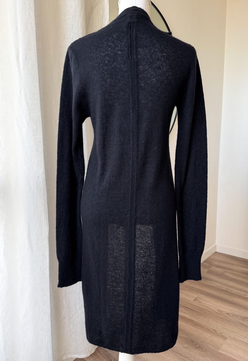 May include: A long, black cardigan sweater with long sleeves. The sweater is made of a soft, textured material and has a simple, elegant design. The back of the sweater is visible, showing a vertical seam.