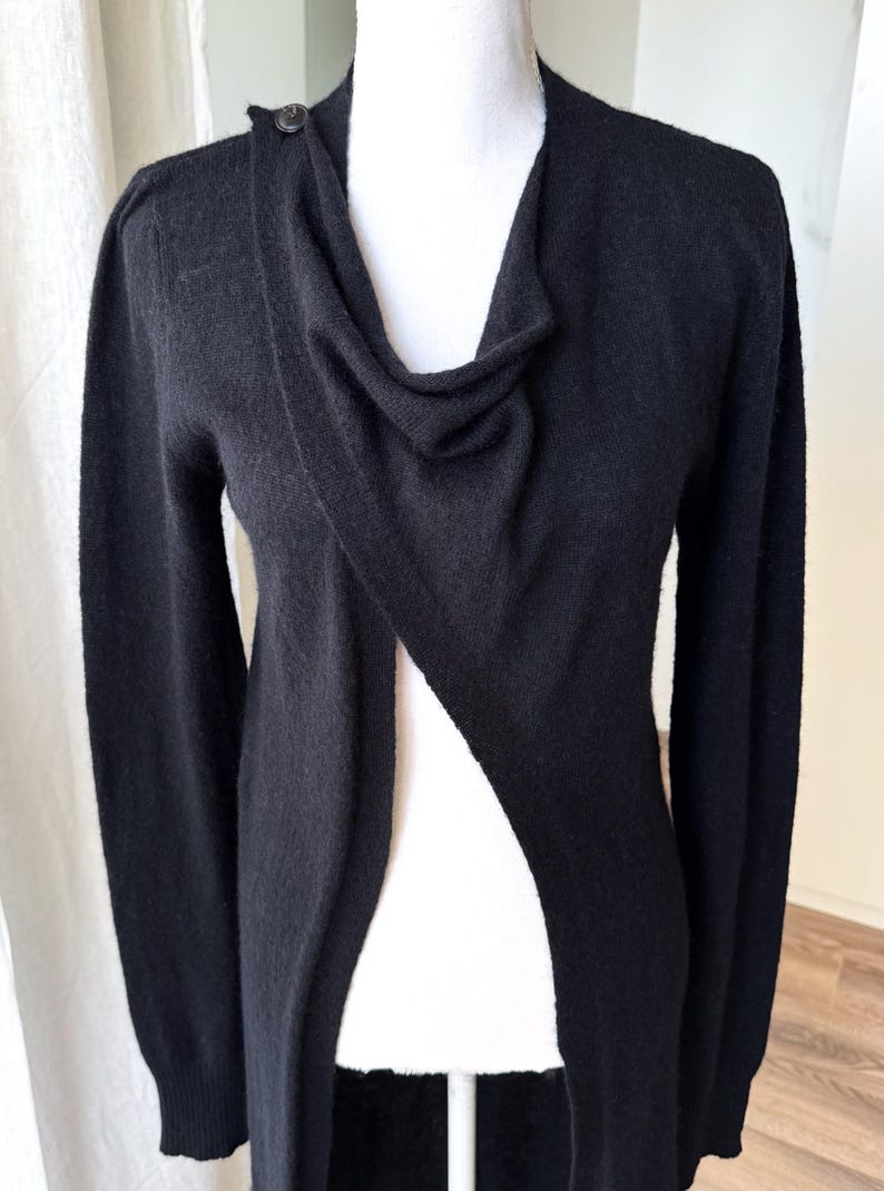 May include: Black long-sleeve cardigan with a draped neckline and a single button closure at the shoulder. The open front design creates a flowing silhouette. The cardigan is made of a soft, knit material.