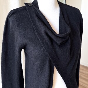 May include: A black cardigan sweater with a draped neckline and a single button closure at the shoulder. The long-sleeved sweater is displayed on a white mannequin.