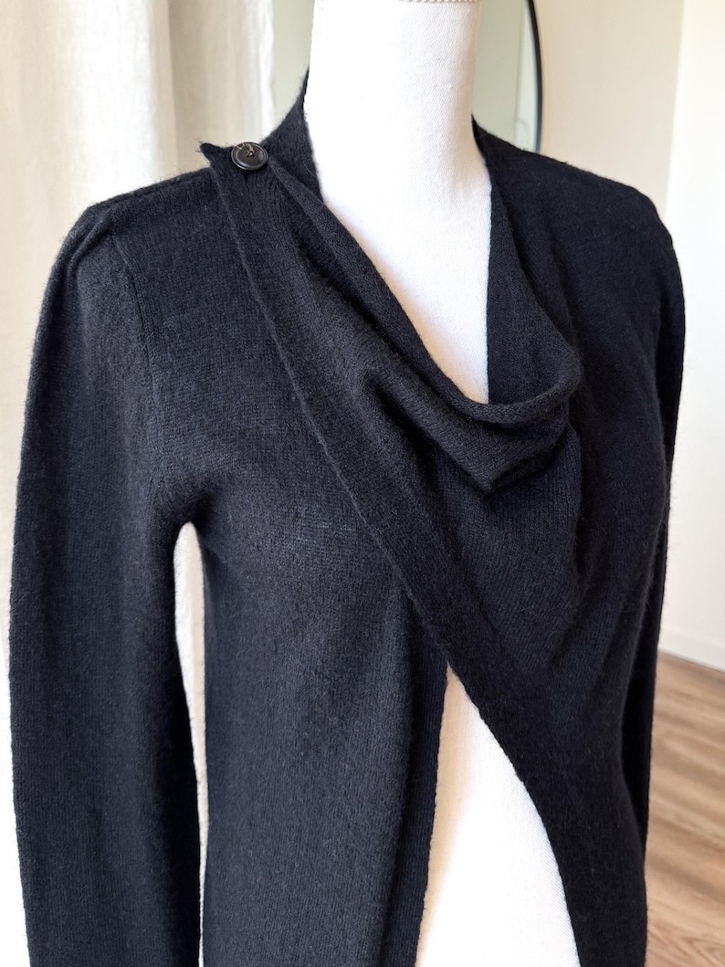 May include: A black cardigan sweater with a draped neckline and a single button closure at the shoulder. The long-sleeved sweater is displayed on a white mannequin.
