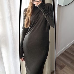 May include: A long-sleeved, dark olive-green dress with a turtleneck. The dress is knee-length and form-fitting, reflecting in a full-length mirror. The person is holding a phone in front of their face.