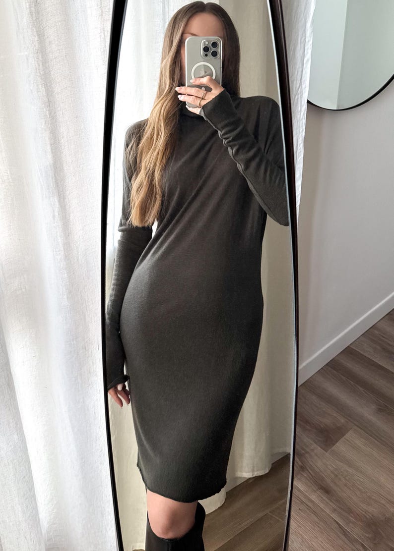 May include: A long-sleeved, dark olive-green dress with a turtleneck. The dress is knee-length and form-fitting, reflecting in a full-length mirror. The person is holding a phone in front of their face.