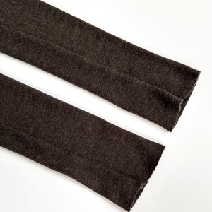 May include: Close-up of two sections of dark brown fabric, possibly sleeves or legs of a garment, against a white background. The fabric has a textured appearance and a visible seam running down the center of each section. The edges are finished.