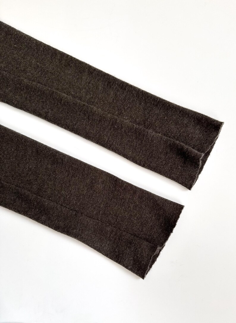 May include: Close-up of two sections of dark brown fabric, possibly sleeves or legs of a garment, against a white background. The fabric has a textured appearance and a visible seam running down the center of each section. The edges are finished.