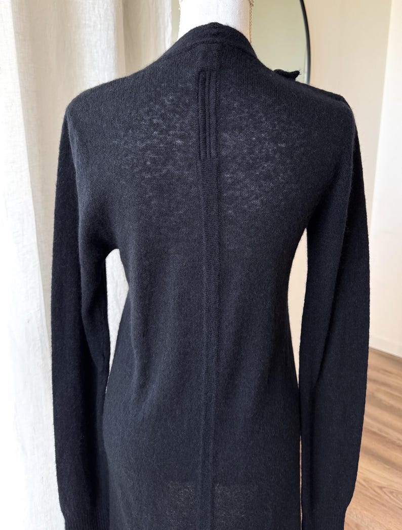 May include: A black long-sleeve cardigan with a V-neck design. The back of the cardigan features a vertical seam detail. The garment is displayed on a mannequin, showcasing its shape and texture. The cardigan is made of a lightweight knit material.