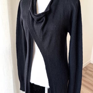 May include: A black cardigan with a draped cowl neckline and long sleeves. The open-front design features a single button closure at the shoulder. The sweater is displayed on a mannequin.