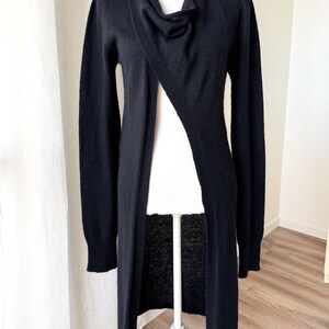 May include: A long, black cardigan with a draped neckline and a single button closure. The open front design reveals the mannequin beneath. The sleeves are long, and the cardigan falls to the ankles.