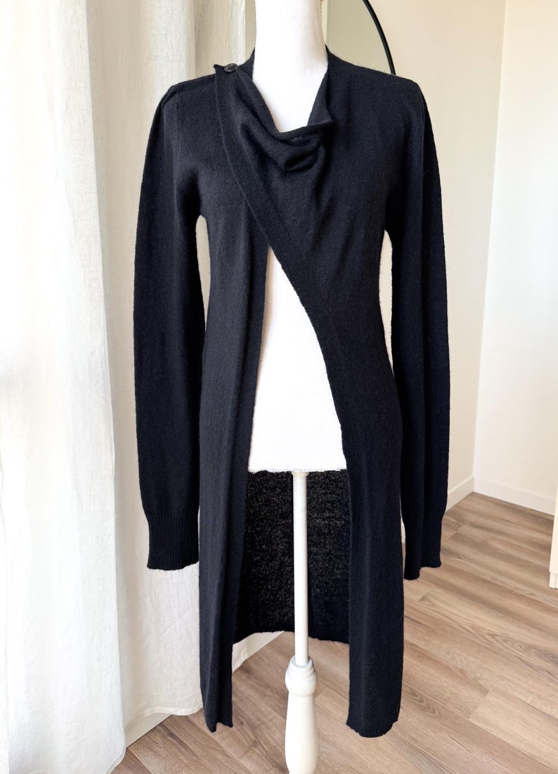 May include: A long, black cardigan with a draped neckline and a single button closure. The open front design reveals the mannequin beneath. The sleeves are long, and the cardigan falls to the ankles.