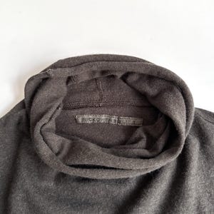 May include: Close-up of a dark gray turtleneck sweater. The sweater has a wide, folded collar and a label with the text "RICK OWENS LICA". The fabric appears to be a soft knit material. The overall style is minimalist and modern.