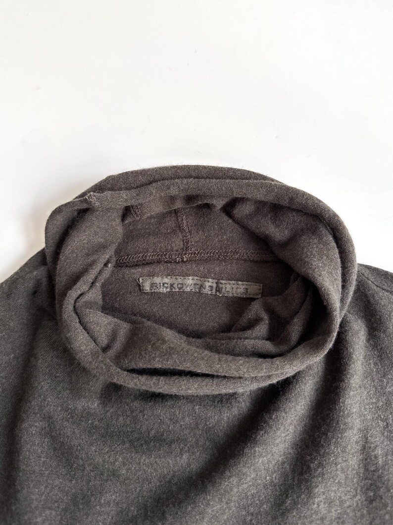 May include: Close-up of a dark gray turtleneck sweater. The sweater has a wide, folded collar and a label with the text "RICK OWENS LICA". The fabric appears to be a soft knit material. The overall style is minimalist and modern.