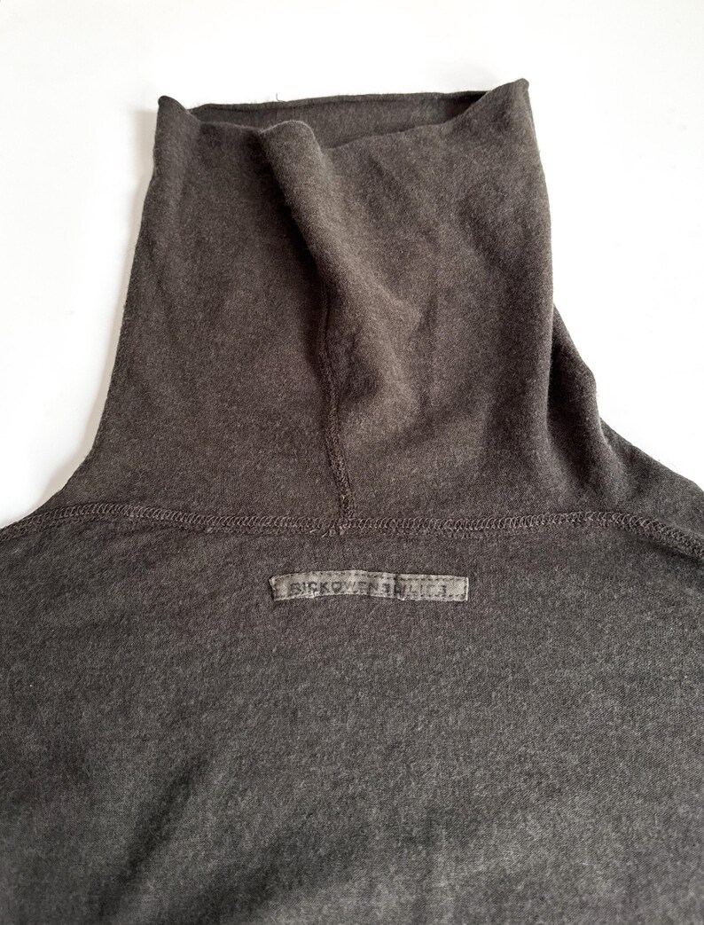 May include: Close-up of a dark gray turtleneck shirt. The shirt has a high, folded collar and a small rectangular label near the neckline with the text "RICKOWENS DRKSHDW". The fabric appears soft and slightly textured.