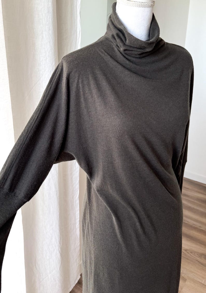 May include: A dark gray long-sleeve dress with a turtleneck collar. The dress has a relaxed fit with long sleeves and a simple, elegant design. The fabric appears to be soft and drapes well. The dress is displayed on a mannequin.