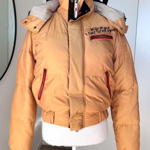 Pode incluir: Casaco puffer bege com capuz. O casaco tem gola alta, zíperes vermelhos e o texto "Mille Miglia race turns legend" no peito. O casaco é exibido em um manequim.