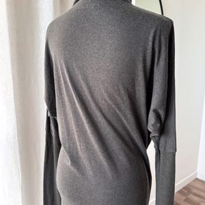May include: A dark gray long-sleeve dress with a cowl neck. The dress is made of a soft, flowing fabric and has long sleeves. The dress is displayed on a mannequin.