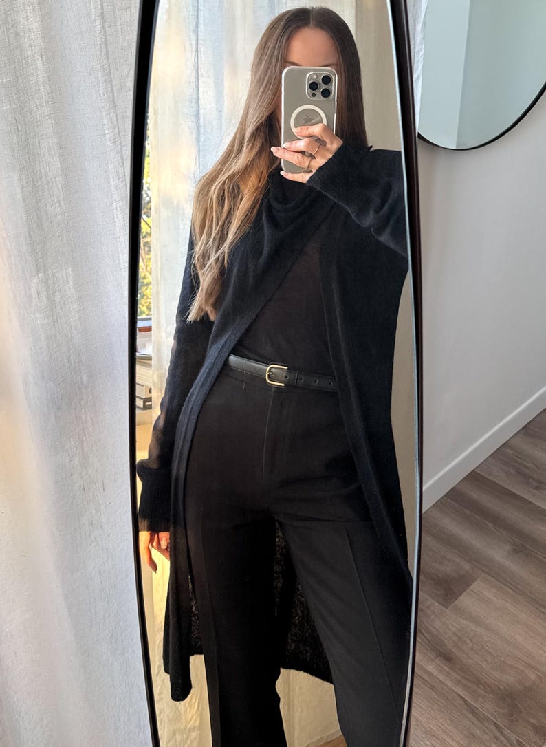 May include: Full-length mirror reflection of a black outfit. The ensemble includes a long, open cardigan, a top, and tailored pants. A thin black belt with a gold buckle cinches the waist. The outfit is monochromatic, creating a sleek, modern look.