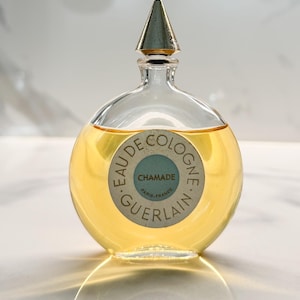 Könnte beinhalten: Eine Vintage-Glasflasche von Chamade Eau de Cologne von Guerlain. Die Flasche ist rund mit einem silbernen Verschluss und einem gelben Etikett mit dem Text "Chamade Paris-France Guerlain" in Schwarz.
