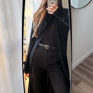May include: Full-length mirror reflection of a black outfit. The outfit includes a long, open cardigan, a top, and tailored pants. A thin black belt with a gold buckle cinches the waist. The overall look is monochromatic and sleek.