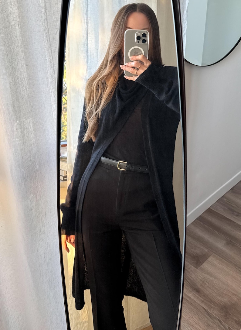May include: Full-length mirror reflection of a black outfit. The outfit includes a long, open cardigan, a top, and tailored pants. A thin black belt with a gold buckle cinches the waist. The overall look is monochromatic and sleek.