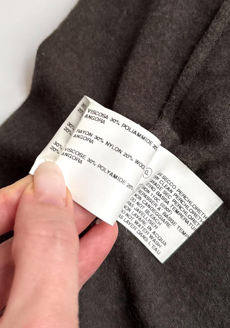 May include: Close-up of a white clothing tag with text in Italian and English. The tag lists fabric composition: Viscose, Polyamide, Rayon, Nylon, Wool, and Angora. Care instructions are also printed on the tag.