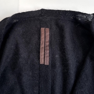 May include: Close-up of a black cardigan. The garment has a soft, textured appearance. A brown label with text is sewn on the back of the collar. The cardigan is made of a dark, possibly wool, material.