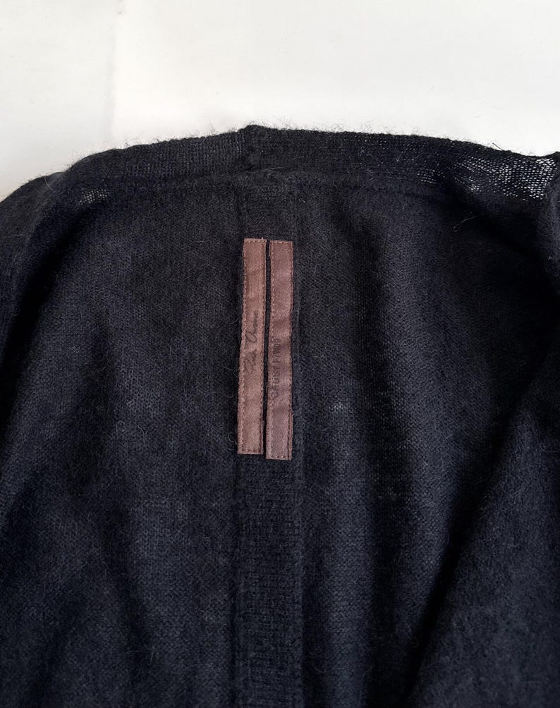 May include: Close-up of a black cardigan. The garment has a soft, textured appearance. A brown label with text is sewn on the back of the collar. The cardigan is made of a dark, possibly wool, material.