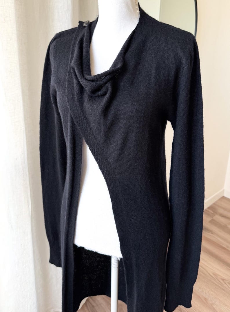 May include: A black cardigan with a draped cowl neckline and long sleeves. The open-front design features a single button closure at the shoulder. The sweater is displayed on a mannequin.