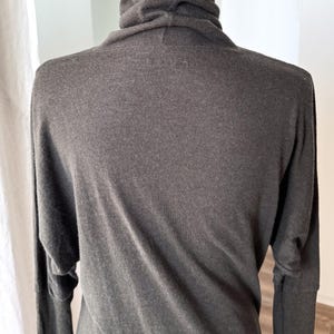 May include: A dark gray long-sleeved top with a cowl neck. The top has a relaxed fit with long sleeves. The fabric appears to be soft and drapes well. The top is displayed on a mannequin.