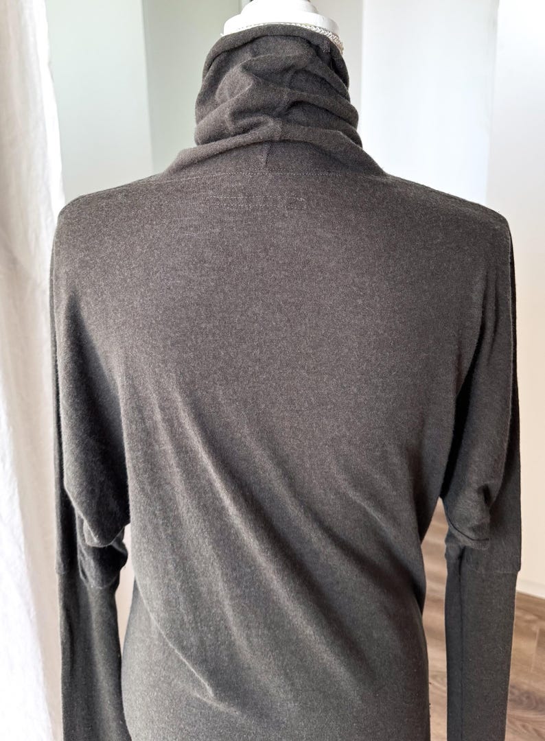 May include: A dark gray long-sleeved top with a cowl neck. The top has a relaxed fit with long sleeves. The fabric appears to be soft and drapes well. The top is displayed on a mannequin.