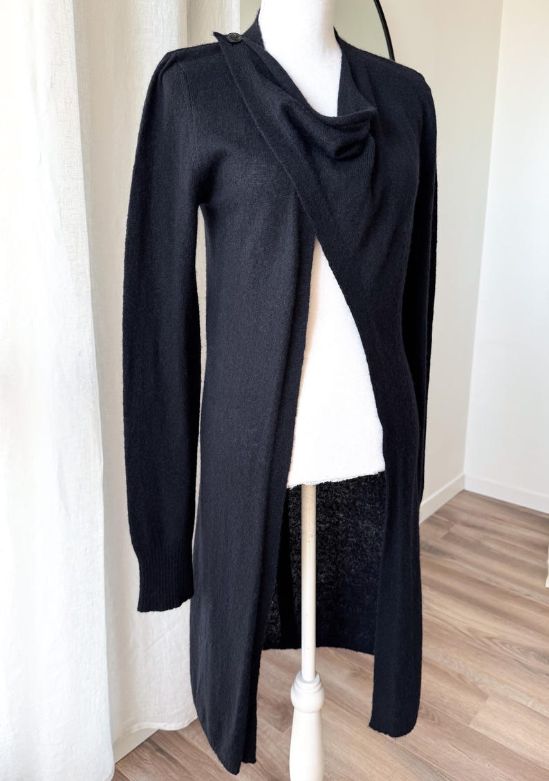 May include: A long, black cardigan with a draped neckline and button closure. The open front design reveals a white garment underneath. The sleeves are long, and the cardigan appears to be made of a soft material. The garment is displayed on a mannequin.
