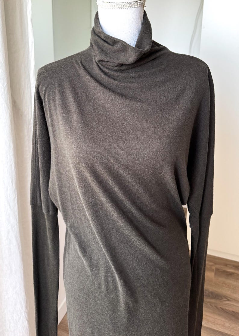 May include: A dark gray long-sleeved top with a cowl neck. The top is made of a soft, flowing fabric and has a relaxed fit. The sleeves are long and the neckline is draped. The top is displayed on a mannequin.