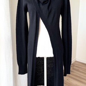 May include: A long, black cardigan sweater with a draped neckline and a single button closure. The sweater is displayed on a mannequin, showcasing its flowing design and long sleeves. The garment is made of a soft material.
