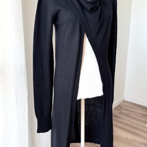May include: A long, black cardigan with a draped neckline and button closure. The open front design reveals a white garment underneath. The sleeves are long, and the cardigan appears to be made of a soft material. The garment is displayed on a mannequin.
