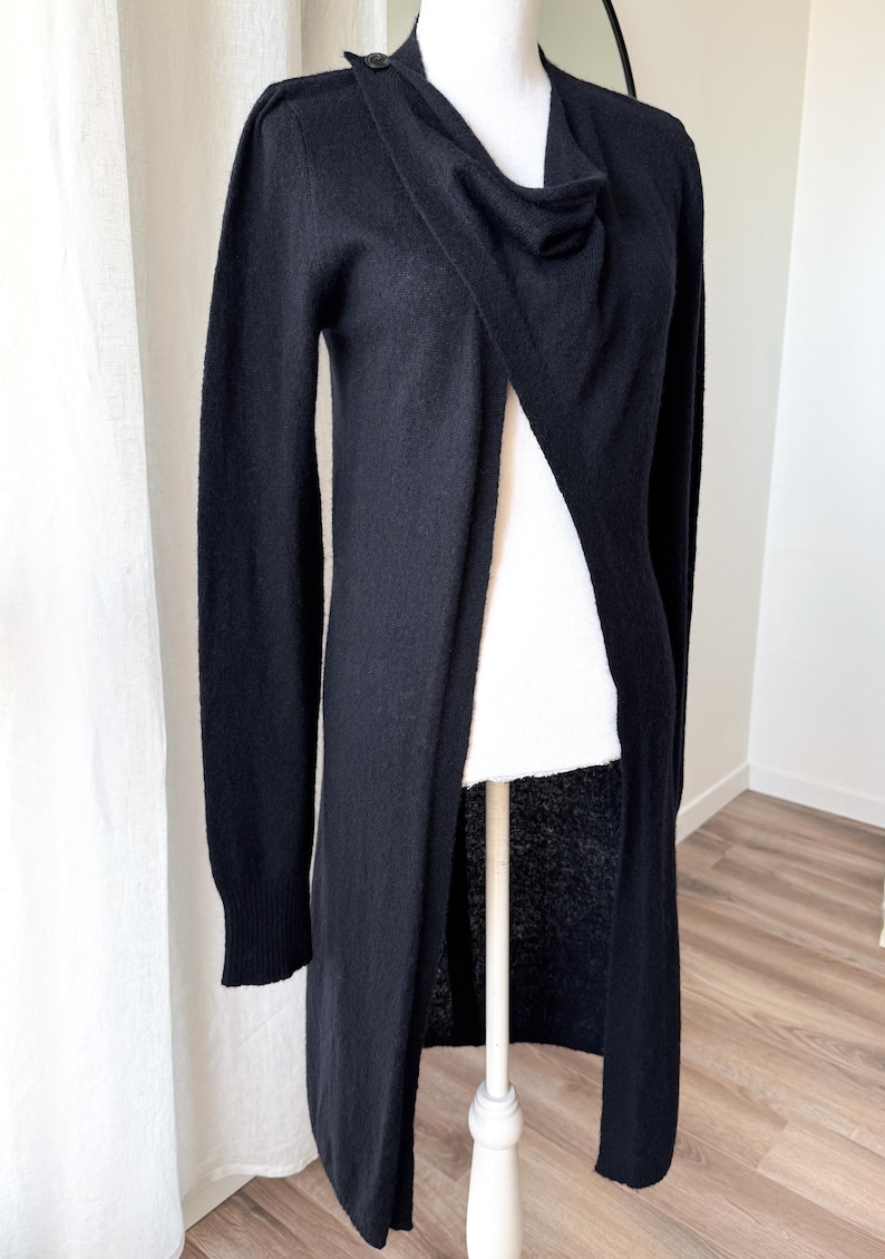 May include: A long, black cardigan with a draped neckline and button closure. The open front design reveals a white garment underneath. The sleeves are long, and the cardigan appears to be made of a soft material. The garment is displayed on a mannequin.