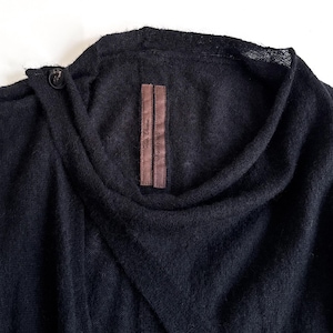 May include: A dark navy blue sweater with a unique asymmetrical design. The sweater features a draped neckline and a single button closure. A brown label with text is sewn on the front. The fabric appears soft and textured.