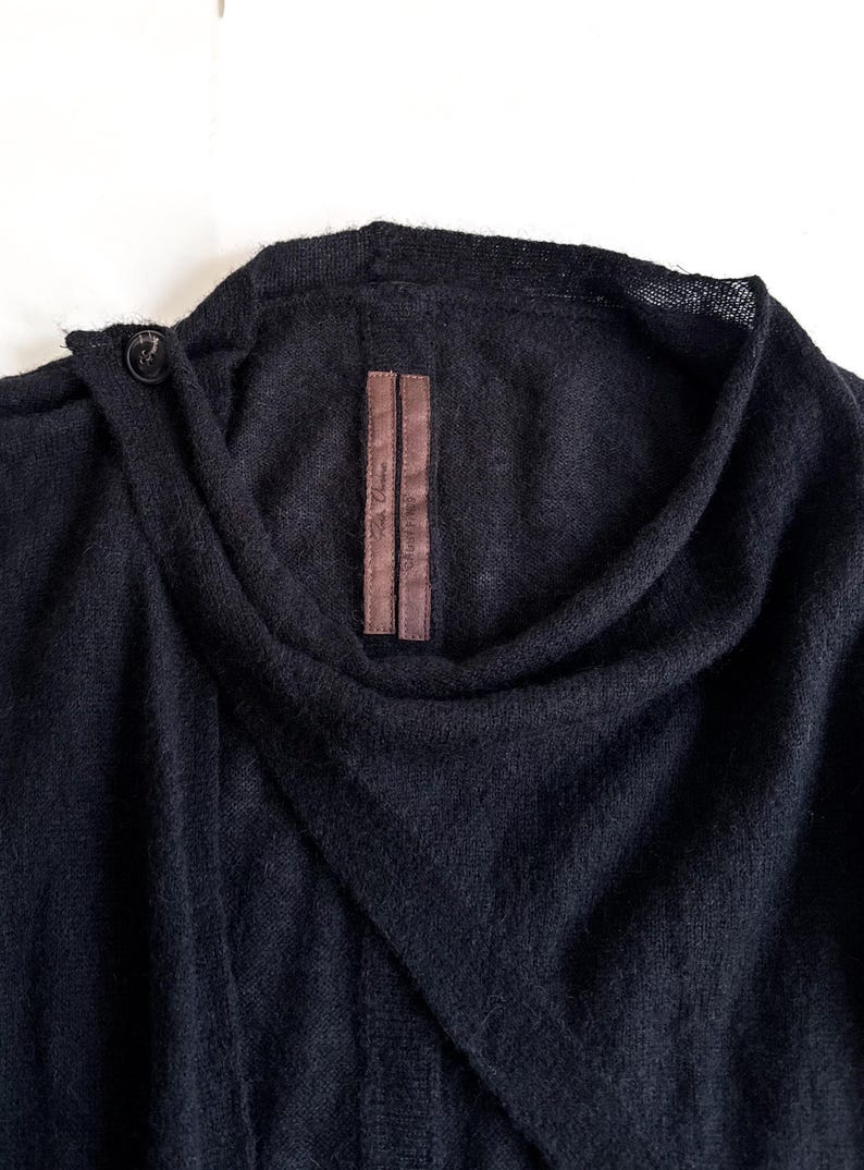 May include: A dark navy blue sweater with a unique asymmetrical design. The sweater features a draped neckline and a single button closure. A brown label with text is sewn on the front. The fabric appears soft and textured.