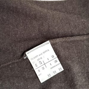 May include: Close-up of dark brown fabric with a white tag attached. The tag reads "OLMAR and MIRTA" and includes size information for ITA, GB, DE, and USA. The fabric has a textured appearance.