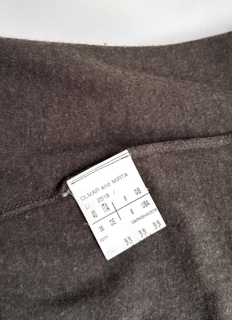 May include: Close-up of dark brown fabric with a white tag attached. The tag reads "OLMAR and MIRTA" and includes size information for ITA, GB, DE, and USA. The fabric has a textured appearance.