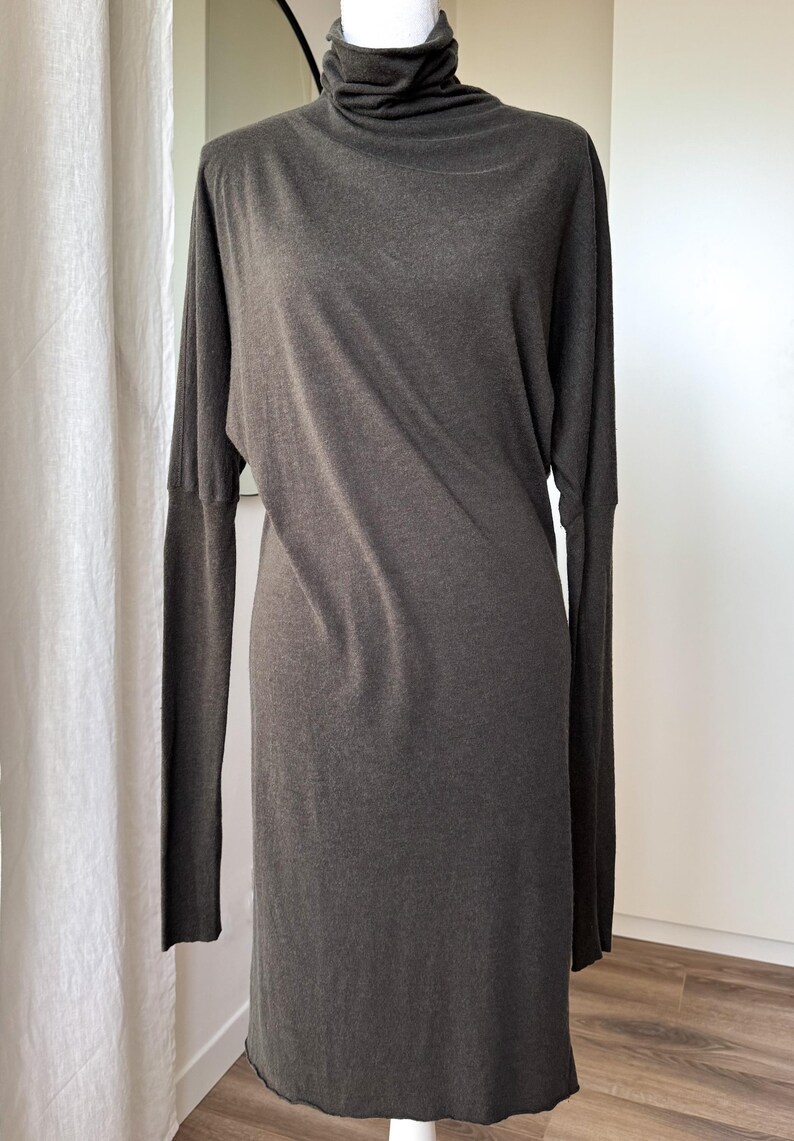 May include: A dark gray, long-sleeved dress with a turtleneck collar. The dress has a simple, streamlined design and falls to the mid-calf. The fabric appears to be a soft knit material, creating a relaxed silhouette.