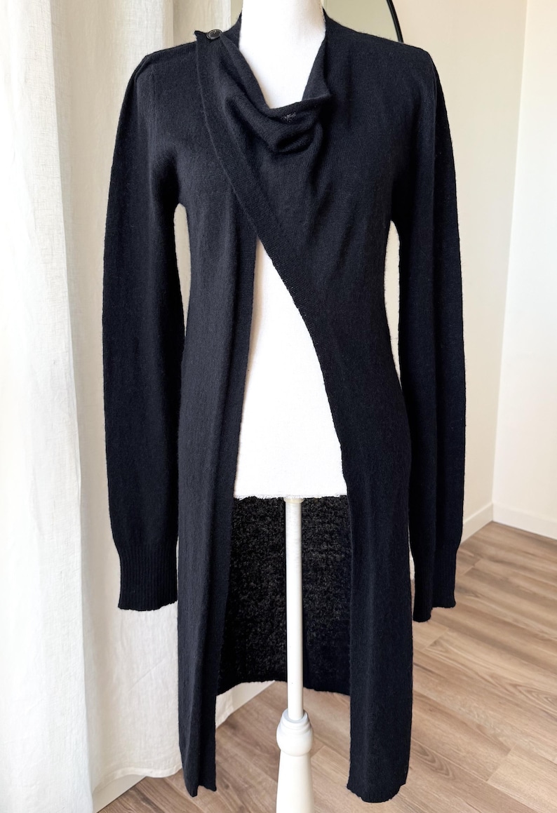 May include: A long, black cardigan with a draped neckline and a single button closure. The open front design reveals a white garment underneath. The sleeves are long, and the cardigan is displayed on a mannequin.