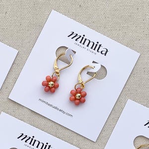 May include: A pair of gold-colored hoop earrings with coral-colored flower charms. The earrings are displayed on white cards with the text "mimita JEWELRY STUDIO" and the website mimitastudio.etsy.com.