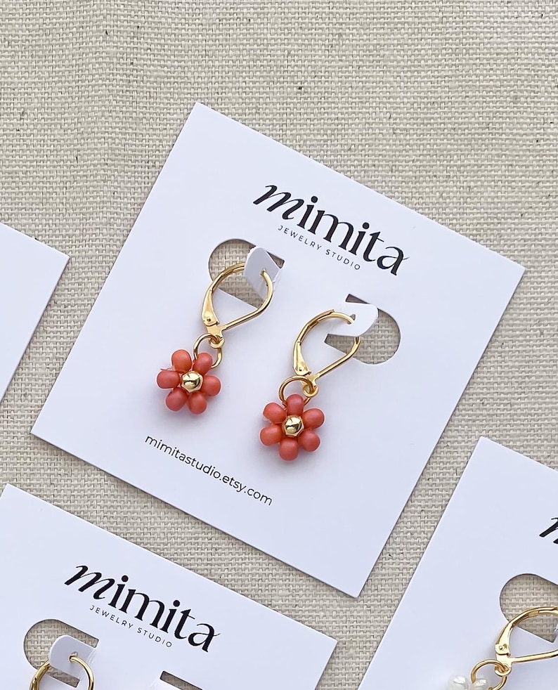 May include: A pair of gold-colored hoop earrings with coral-colored flower charms. The earrings are displayed on white cards with the text "mimita JEWELRY STUDIO" and the website mimitastudio.etsy.com.