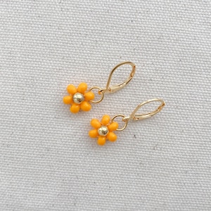 May include: A pair of gold-colored hoop earrings, each featuring a small, orange daisy pendant with a gold-colored center. The earrings are displayed on a textured, off-white surface.
