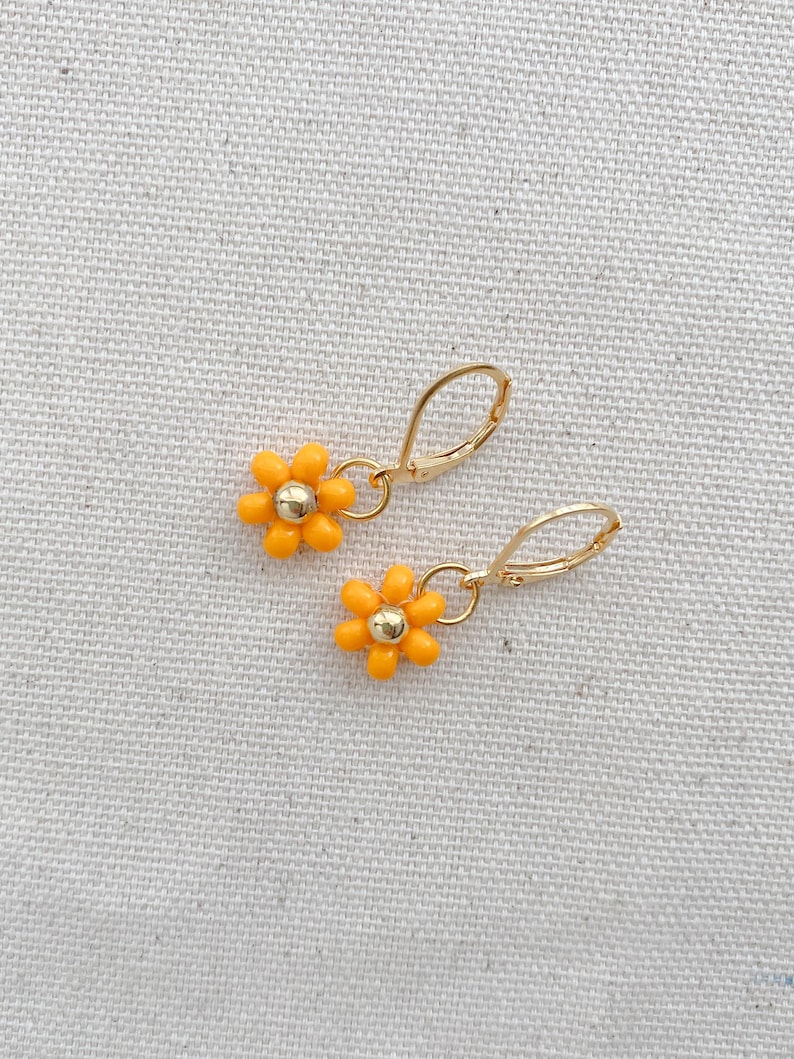May include: A pair of gold-colored hoop earrings, each featuring a small, orange daisy pendant with a gold-colored center. The earrings are displayed on a textured, off-white surface.